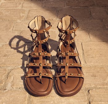 Sandalias gladiador de cuero marrón con tachuelas doradas, múltiples correas y cierres en el tobillo sobre un suelo de baldosas de piedra a la luz del sol.
