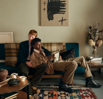 Δύο άντρες κάθονται σε έναν τυρκουάζ καναπέ με ριγέ ριχτάρι. Ο ένας διαβάζει ένα βιβλίο, φορώντας ένα μπεζ σακάκι· ο άλλος σκύβει προς τα πίσω. Κοντά τους υπάρχει ένα μαύρο τσαγιέρα και φλυτζάνια.