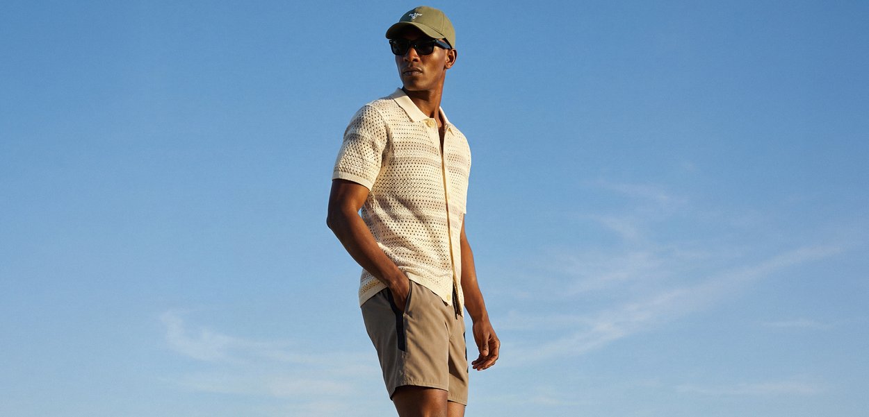 Tricou polo bej cu mâneci scurte și model texturat, asortat cu pantaloni scurți bej. Bărbatul poartă ochelari de soare și o șapcă verde, pe fundal albastru de cer.