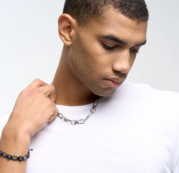 Zilveren metalen ketting met slot, gedragen over een eenvoudig wit shirt. Kenmerkt zich door gladde schakels en een bijpassende zwarte armband.