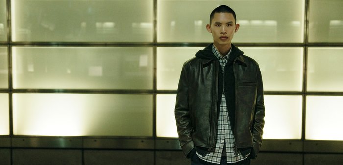 Brown leather jacket over a dark hoodie, paired with a checked shirt. Smooth texture with pockets. Neutral background with soft lighting.