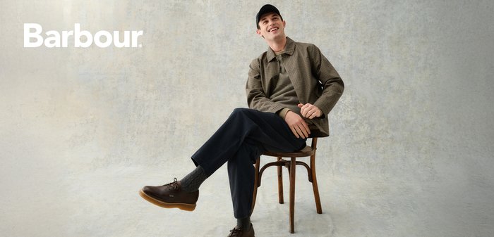 Braun kariertes Jacket über einem grauen Pullover, marineblaue Hose, graue Socken und braune Schuhe. Model sitzt auf einem Holzstuhl vor einem strukturierten Hintergrund.