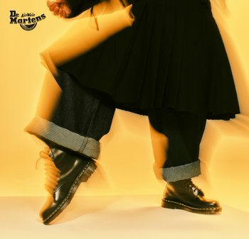 Botas de cuero negras hasta el tobillo con costuras amarillas, combinadas con jeans de mezclilla azules remangados en los puños, sobre un fondo amarillo.