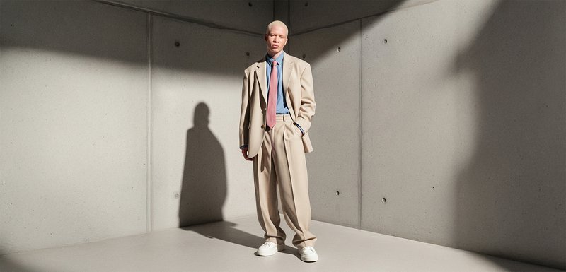 Costume beige surdimensionné avec une chemise bleu clair, une cravate rose et des baskets blanches, présentant un design ajusté et texturé sur fond de béton.