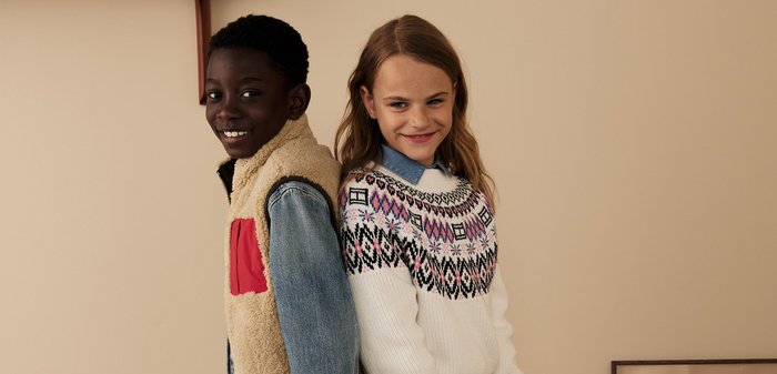 Zwei Kinder stehen Rücken an Rücken. Eines trägt eine beigefarbene Fleeceweste mit einer roten Tasche und ein Jeanshemd; das andere hat einen weißen gemusterten Pullover mit schwarzen Akzenten.