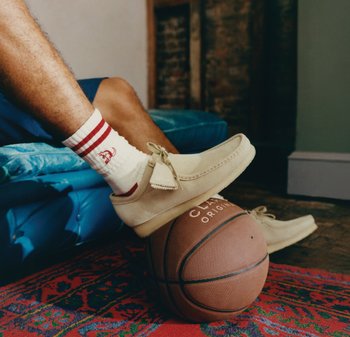 Personne portant des mocassins en daim beige et des chaussettes blanches à rayures rouges repose le pied sur un ballon de basket posé sur un tapis rouge à motifs.
