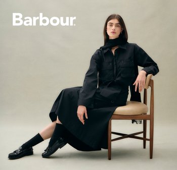 Schwarzes langes Hemdkleid mit zwei frontalen Taschen, über einem schwarzen Rock getragen. Kombiniert mit schwarzen Socken und Slippern. Natürlicher Holzstuhl.