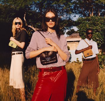 Trois femmes portant des lunettes de soleil, tenant des sacs à main de créateurs, debout dans l'herbe en plein air avec des arbres et un bâtiment en arrière-plan.