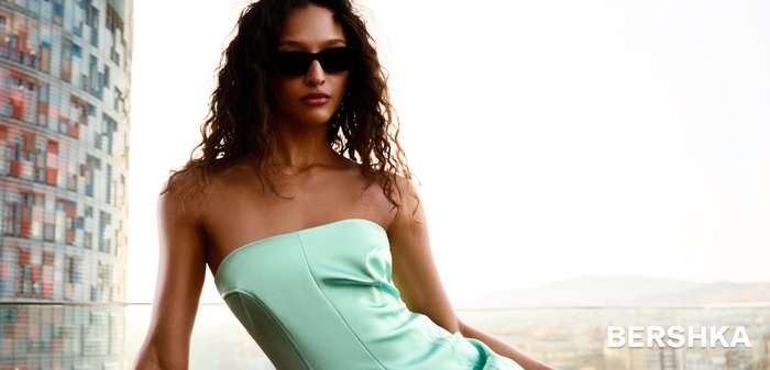 Femeie cu ochelari de soare purtând o rochie fără bretele verde mentă, așezată pe o margine, cu peisaj urban în fundal.