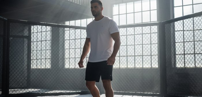 Man wearing white t-shirt, black shorts, and sneakers standing in a sunlit indoor cage with large windows and shadows on the floor.