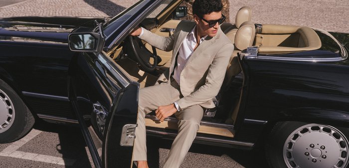 Man in beige suit and loafers sitting in open door of black vintage convertible car, looking to the side wearing sunglasses.