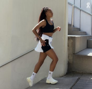 Zwarte sportbeha, zwarte shorts met logo, wit knoopshirt, witte sokken met logo en lichtgekleurde sportschoenen. Trappen op de achtergrond.