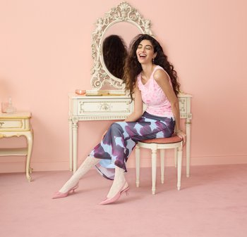 Giovane donna dai capelli ricci seduta su uno sgabello accanto a uno specchio da toeletta ornato, indossa un top rosa, una gonna a fantasia e tacchi rosa in una stanza dai toni pastello.
