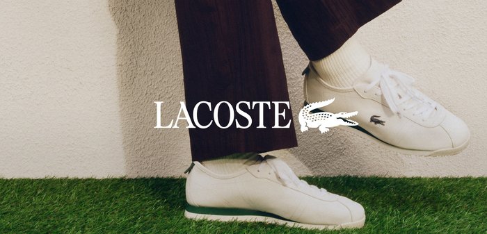 Person sitzt auf einer weissen Kante in der Nähe von Wasser, trägt dunkle Hosen, weisse Socken, weisse Lacoste-Sneakers, mit einem Maschendrahtzaun im Hintergrund.