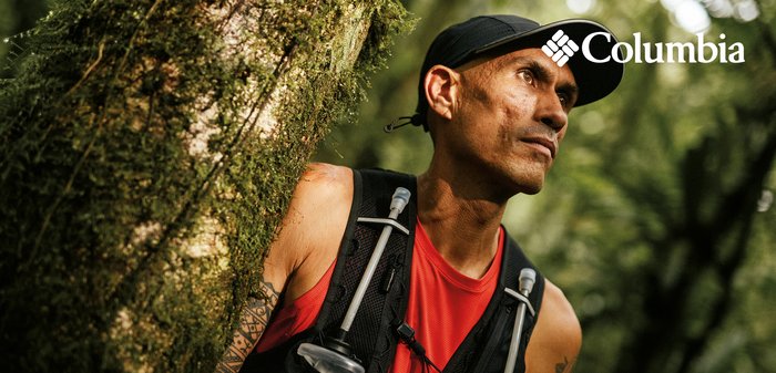 Athlete in red shirt and black cap wearing hydration vest stands by mossy tree in forest, looking intently ahead.