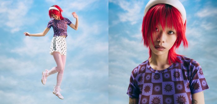 Personne aux cheveux rouge vif portant un béret blanc, une chemise violette à motifs, un short à pois et des chaussures de ballet, posant devant un ciel nuageux.