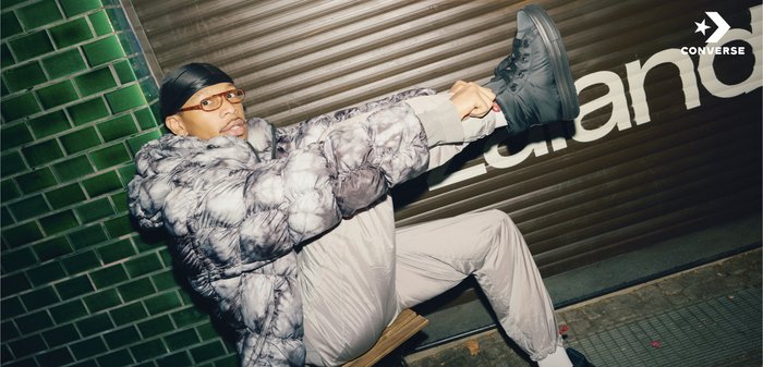 Baskets montantes Converse noires, veste matelassée grise avec un motif tie-dye, pantalon gris, assis sur une chaise devant un mur de carreaux verts.