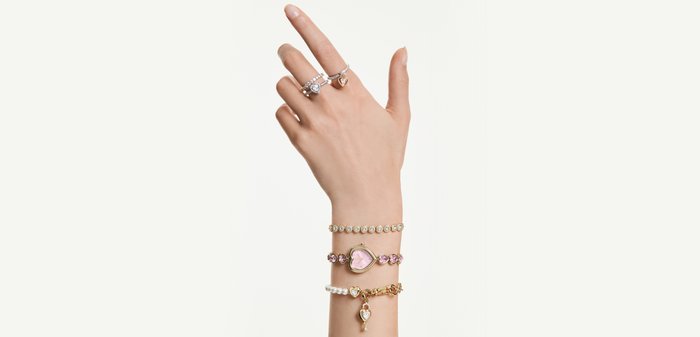 Hand mit Ringen an drei Fingern, die drei Armbänder trägt: Perlenarmband, rosa herzförmiger Anhänger und ein goldenes Armband mit hängendem Herzanhänger.