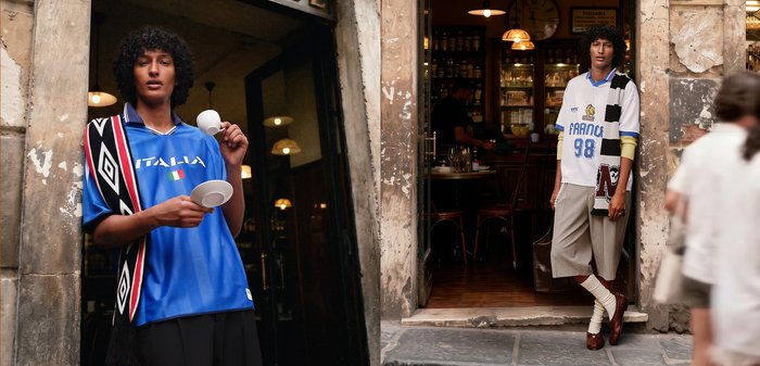 Twee mannen met krullend haar die oversized sporttruien dragen, staan buiten een café in een stenen straat, één houdt een kop en schotel vast.