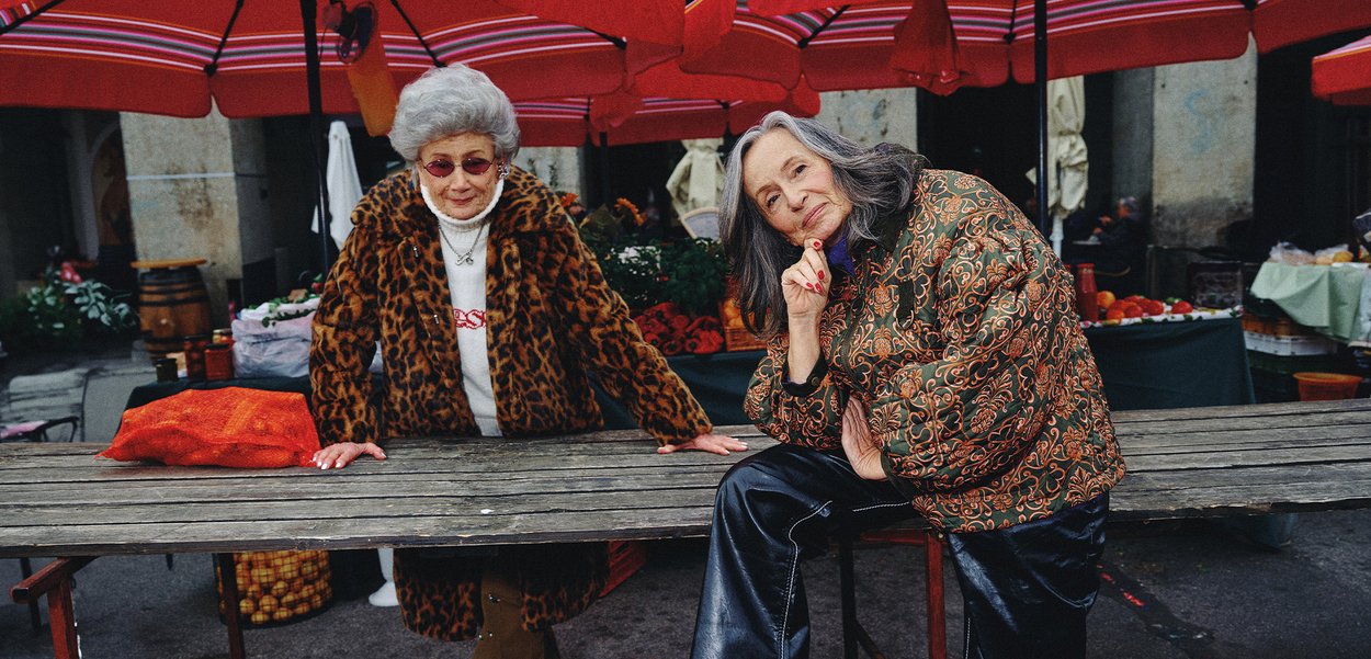 Dvije žene za tržnim stolom: jedna u kaputu sa uzorkom leoparda, a druga u zelenoj jakni s uzorkom. Crveni kišobrani pružaju sjenu.