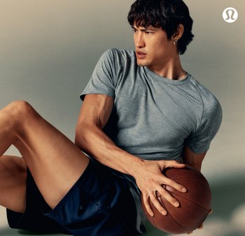 Modello maschile atletico in una maglietta grigia a maniche corte con texture e pantaloni corti scuri, che tiene un pallone da basket marrone, seduto con un'espressione concentrata.