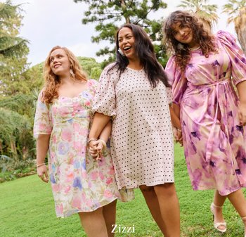 Tre kvinder smilende og holder hinandens hænder, gående på græs; iført farverige blomster- og polkaprikkede kjoler i en udendørs parkscene.
