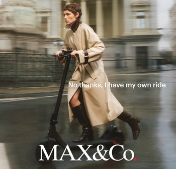 Czarna elektryczna hulajnoga o eleganckim designie, matowym wykończeniu i czerwonych akcentach. Osoba jedzie ubrana w beżowy płaszcz i ciemne buty.