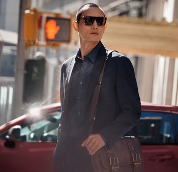 Mężczyzna w ciemnym żakiecie na dżinsowej koszuli, w okularach przeciwsłonecznych, niosący brązową skórzaną torbę na ramię z złotymi akcentami.