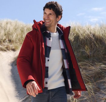 Rote isolierte Jacke mit Reißverschluss und großer Kapuze, über einem weißen strukturierten Pullover und einem karierten Hemd. Blaue Jeans und Strandhintergrund.