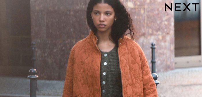 Mladá žena kráčí po městské ulici, má na sobě oranžovou prošívanou bundu, zelený pletený top, hnědou sukni, drží kelímek kávy a tašku.