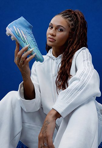 Junge Frau in weißer Sportbekleidung, die einen blauen Fußballschuh mit mehrfarbigen Akzenten vor einem einfarbigen blauen Hintergrund hält.