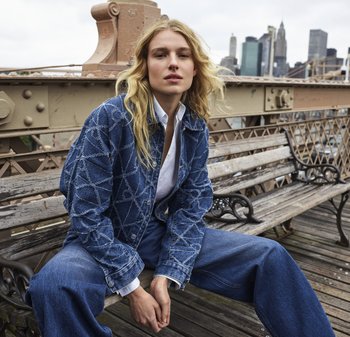 Giacca di jeans con motivo a diamante, colore azzurro chiaro e bottoni argentati. Abbinata a jeans blu larghi e una camicia bianca. Sfondo urbano.