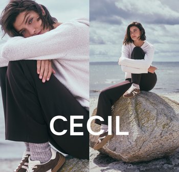 Helles beiges Strickpullover, schwarze Hose, braune Sneaker mit weißen Akzenten, sitzt auf einem Felsen am Strand unter einem bewölkten Himmel.