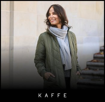 Groene doorgestikte jas, grijze sjaal, donkere trui, staand tegen een stenen muur, met een lichte glimlach en lang haar, handen in de zakken.