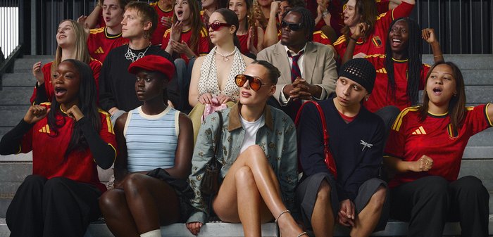 Groupe diversifié de jeunes assis sur les gradins, certains portant des maillots de sport rouges, engagés et encourageant lors d’un événement.
