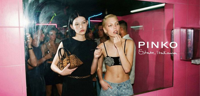 Dwie kobiety w klubie: jedna o czarnych włosach trzyma torebkę w panterkę, druga o krótkich blond włosach nakłada pomadkę.