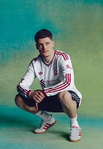 Junger Mann hockt mit verschränkten Händen, trägt weiß-rotes Liverpool-Fußballtrikot, schwarze Shorts und weiße Sneakers vor grünem Hintergrund.