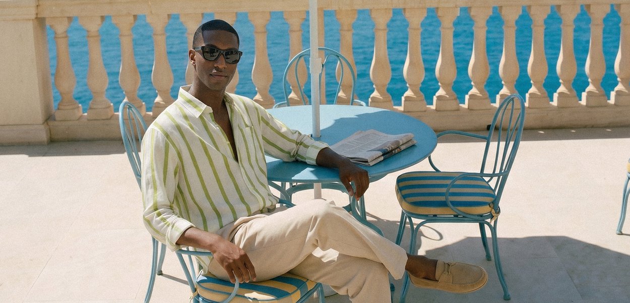 Homme en chemise rayée et lunettes de soleil assis à une table bleue extérieure avec un journal, terrasse en bord de mer avec balustrade en pierre en arrière-plan.