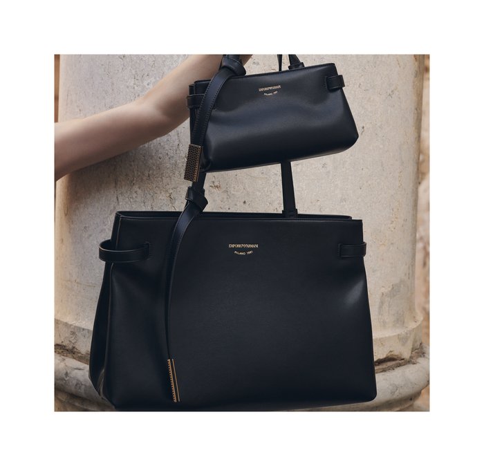 Due borse a tracolla Emporio Armani nere in pelle, una più piccola tenuta sopra una più grande, con uno sfondo di colonne in pietra chiara.