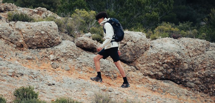 Young person wearing a backpack hikes down a rocky trail surrounded by green shrubs and mountainous terrain.