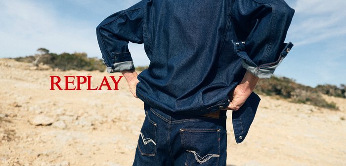Personne portant une veste en jean bleu foncé et un jean avec des coutures blanches, les mains sur les hanches, debout à l'extérieur sur un sol sablonneux.