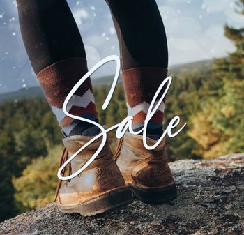 Braune Lederstiefel auf einer felsigen Oberfläche, dazu braune und gemusterte Socken mit roten, blauen und weißen Akzenten, umgeben von einem verschwommenen natürlichen Hintergrund.