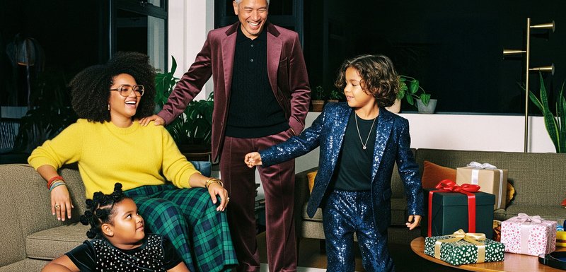 Groupe de quatre personnes en tenues variées, portant un pull jaune, un costume bordeaux et un costume bleu scintillant, avec des cadeaux à proximité.