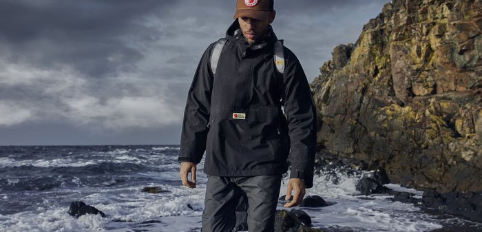 Schwarze wasserdichte Jacke mit einer Fronttasche, verstellbare Kapuze und grauen Hosen. Der Hintergrund zeigt eine felsige Küstenlinie und Meereswellen.