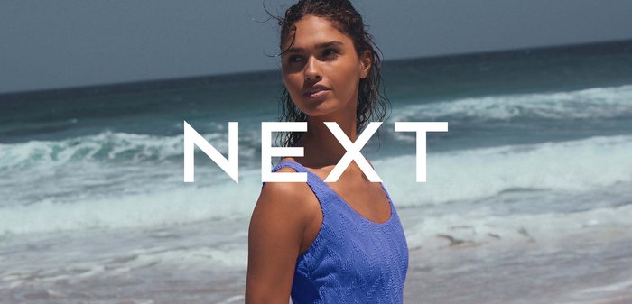 Mujer con un bañador de una pieza azul texturizado caminando por una playa de arena cerca de las olas del océano bajo un cielo despejado con la superposición del texto "NEXT".