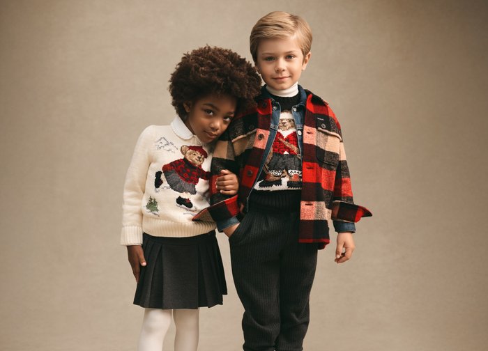 Zwei Kinder stehen nah beieinander, eines legt den Kopf auf die Schulter des anderen und trägt Winterpullover mit Bärenmuster sowie eine karierte Jacke.