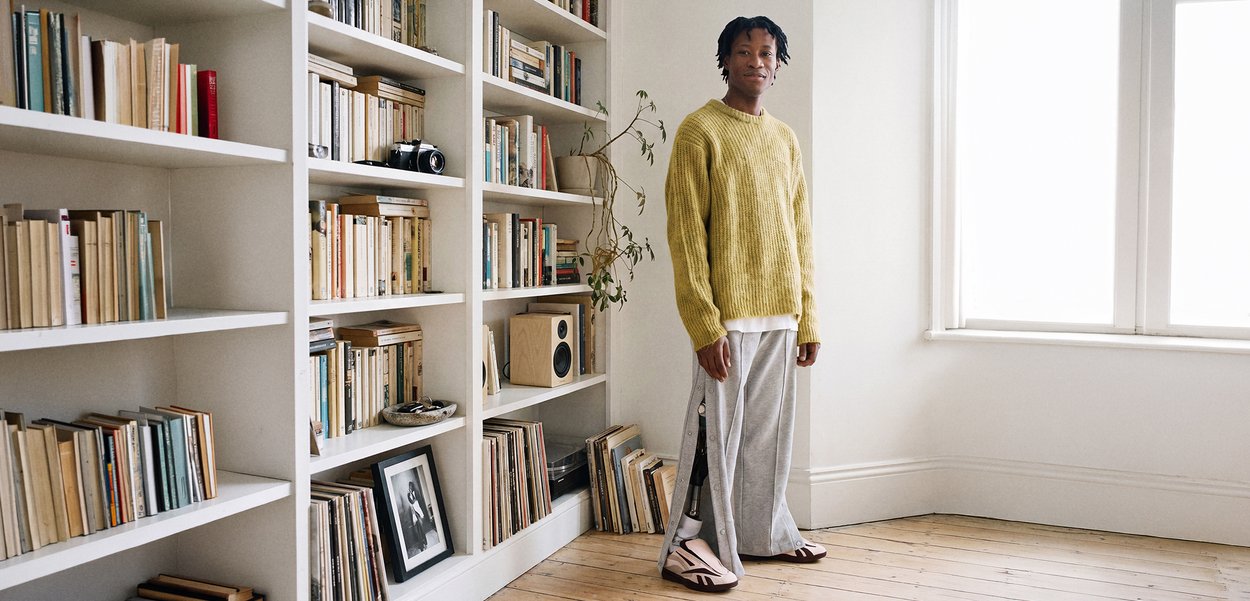 Person wearing a yellow sweater and gray pants stands near white bookshelves filled with books and a speaker in a bright room with wooden floor.