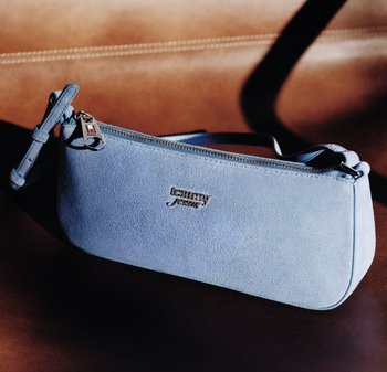 Bolso de mano Tommy Jeans de gamuza azul claro con cremallera y logo metálico, apoyado sobre una superficie de cuero marrón.