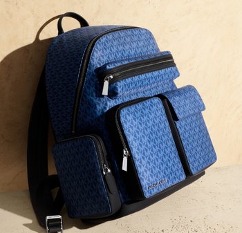 Mochila azul de Michael Kors con múltiples bolsillos con cremallera y solapa, patrón de monograma y correas ajustables para los hombros apoyada contra una pared beige.