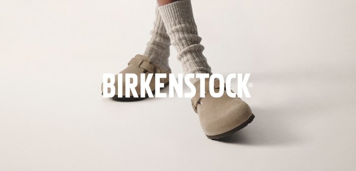 Beige Wildlederclogs mit zwei verstellbaren Riemen, kombiniert mit hellgrauen ribbeligen Socken. Auf einer schwarzen Laufsohle vor einem hellen Hintergrund.